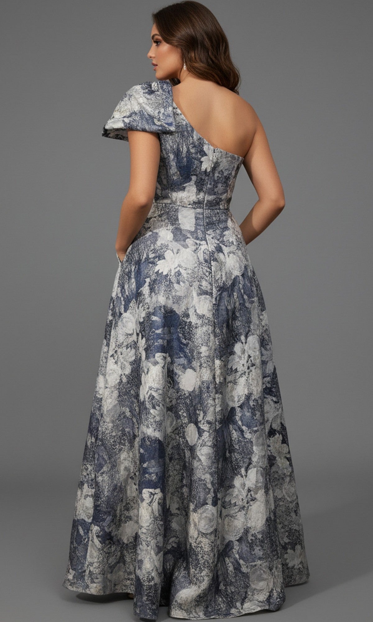 Woman wearing the Jovani 25663 long formal dress, a blue and white floral one-shoulder gown with an elegant a-line skirt, standing with her back to the camera against a plain gray background.
